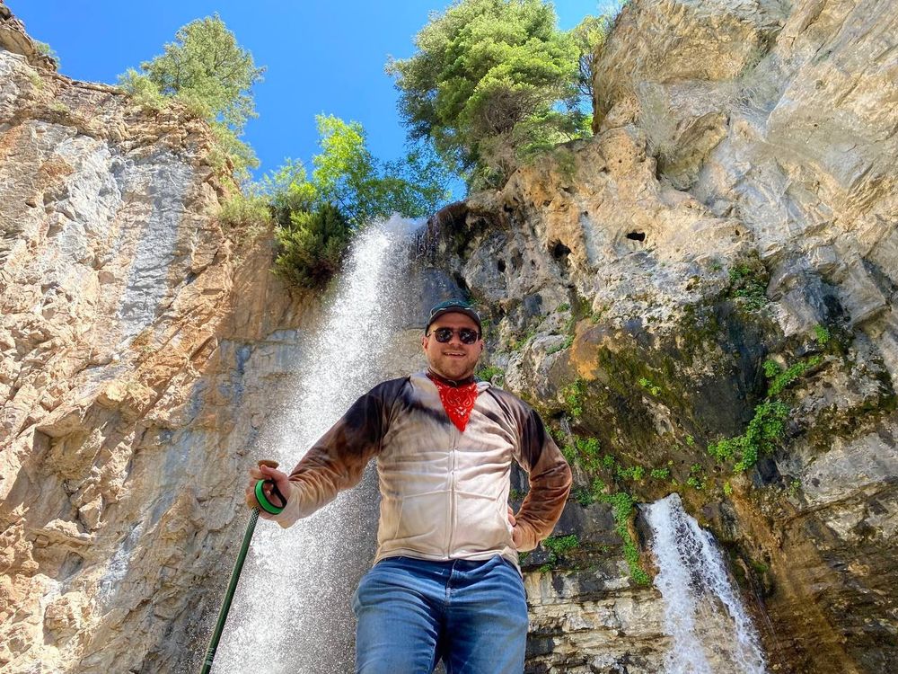A figure in a coyote-print hoodie, wearing a red bandanna, stands over the camera. Behind him, a tall water fall tumbles over a cliff made of yellow-white rock, with a few trees standing above the cliff.