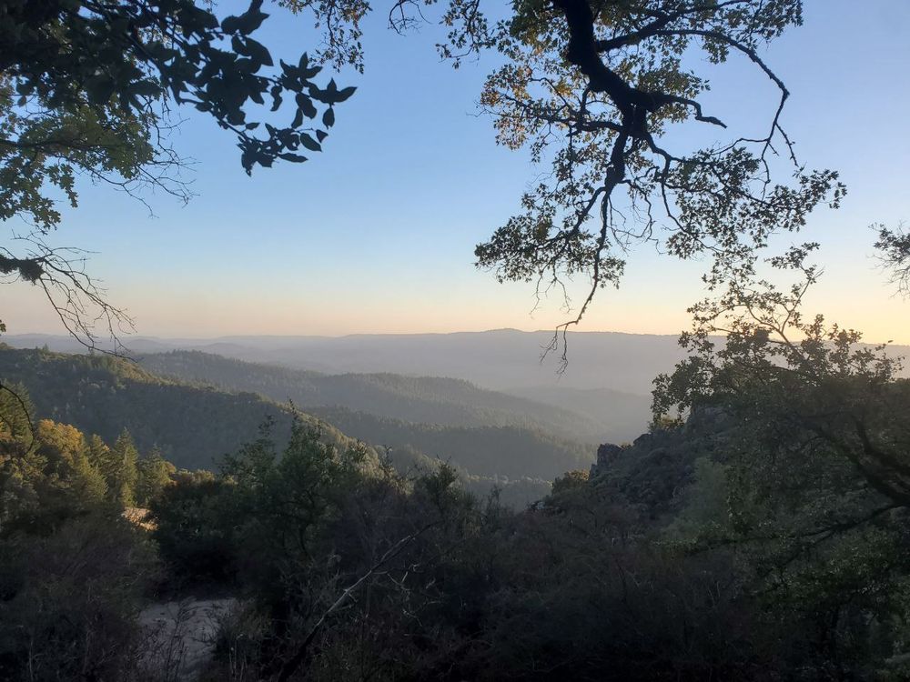 A vista overlooking several mountains, each getting hazier as they recede into the distance. The sky is completely clear, with some orange on the horizon, the sun apparently nearing sunset. The mountains are covered with green trees, too thick to be noticed save their color.
