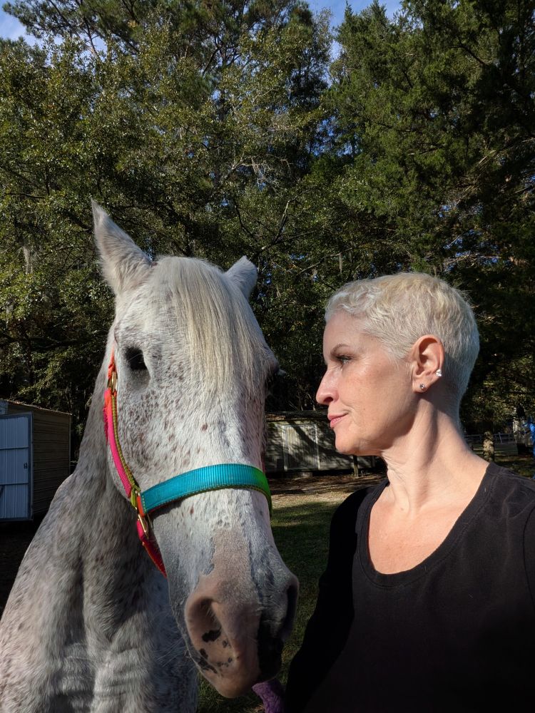 A gray horse with one eye wears a rainbow halter. A woman with short blonde hair gives him a wry look