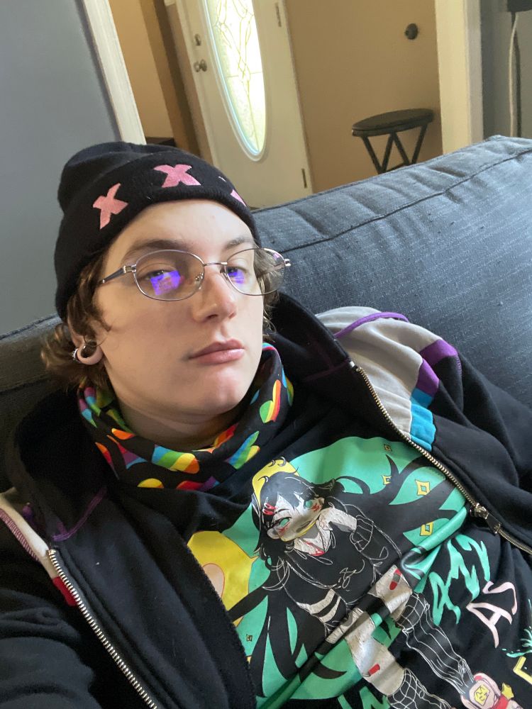 Reiliyana is wearing her black knit cap with “XXX” across the front, a black bandana with rainbow hearts pattern tucked into their black T-shirt with a graphic of a fem-presenting person sticking their toungue out, and a zip-up hoodie. They’re sitting on the couch glaring at you through the camera in a lesbian manner.