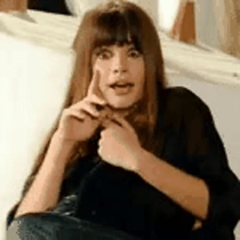 a woman with long hair and bangs is sitting on a couch making a funny face .