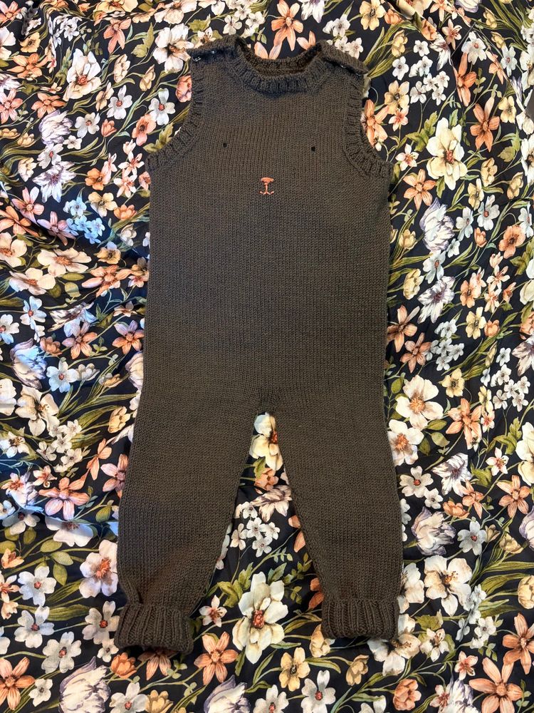 A brown knit onesie. It has legs with ribbed cuffs and no sleeves, though the top is also ribbed all the way across. In the center of the chest is a set of black eyes and small pink nose and mouth embroidered to look like a teddy bear. The onesie is on a pink and black floral duvet for presentation. 
