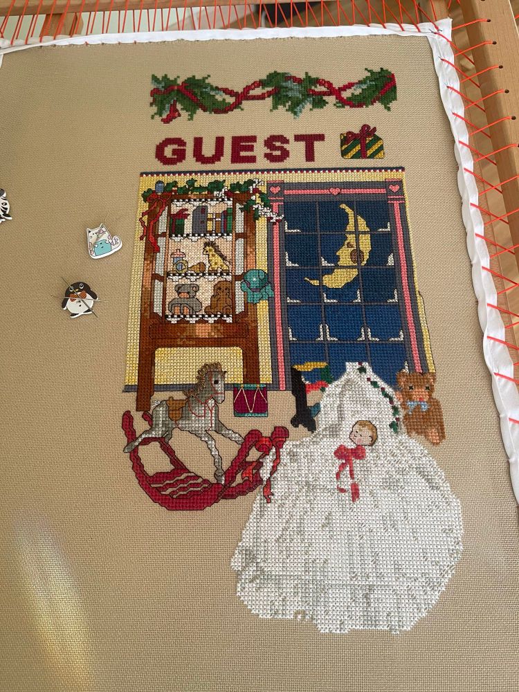 Partially complete Christmas stocking on a dressed slate showing a rocking horse, yellow moon in an icy window, toys on a shelf, and a baby doll in a crib with a white blanket. The top of the stocking has the name “Guest” with a garland header. 