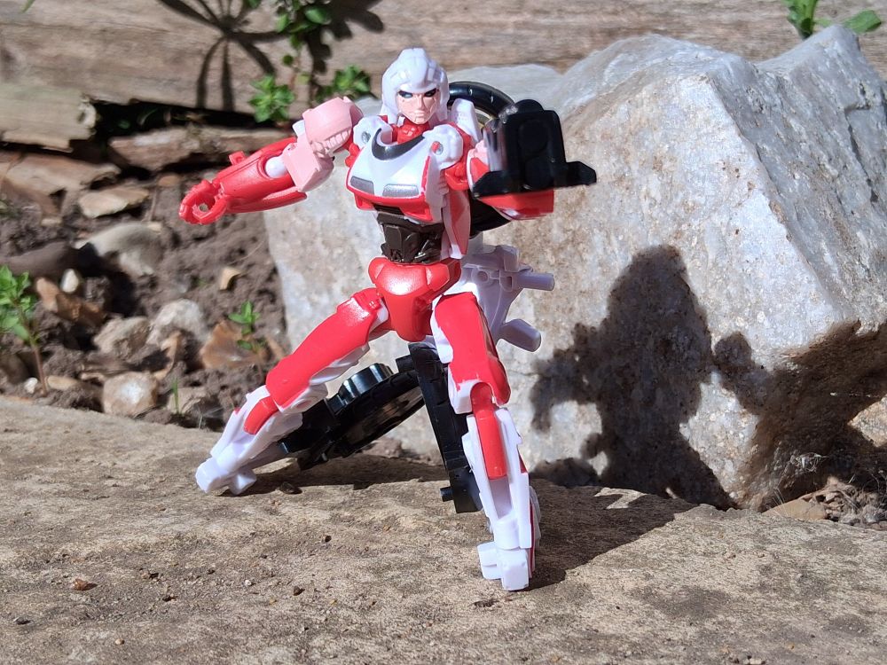 Arcee from Transformers Rise of the Beasts posed outdoors with her gun aiming at the camera. There's a large rock, and some small plants behind her.