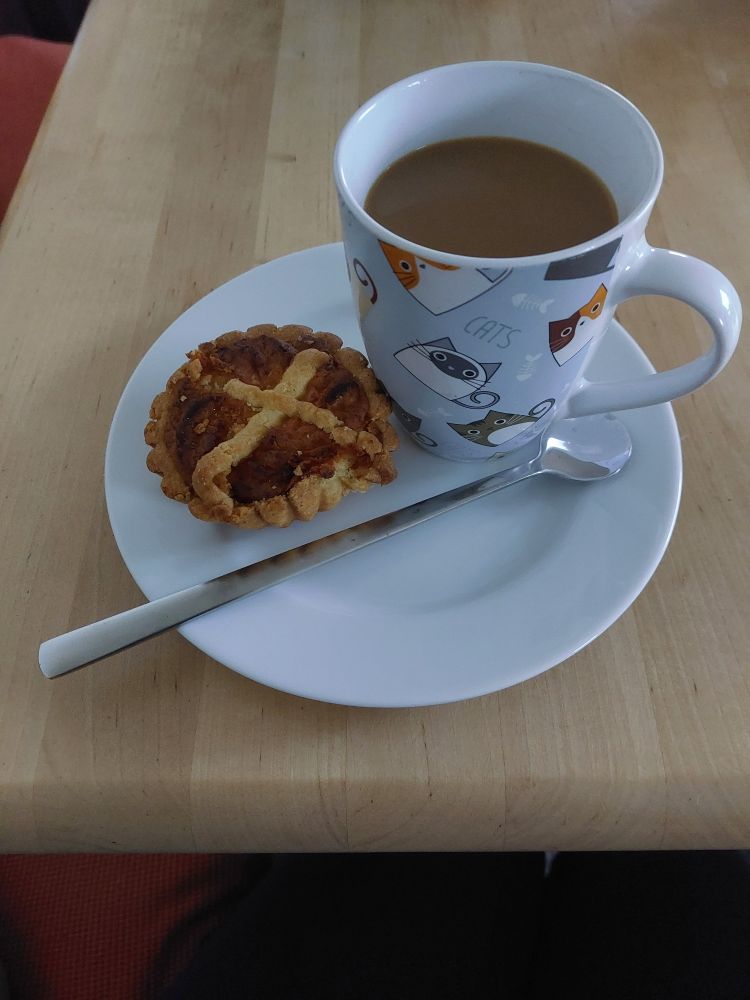 Foto einer sog. Lärche auf einem weißen Teller. Auf dem Teller steht auch eine Tasse mit Kaffee, der heute einen Schuss Milch enthält. Daneben liegt ein langstieliger Löffel.