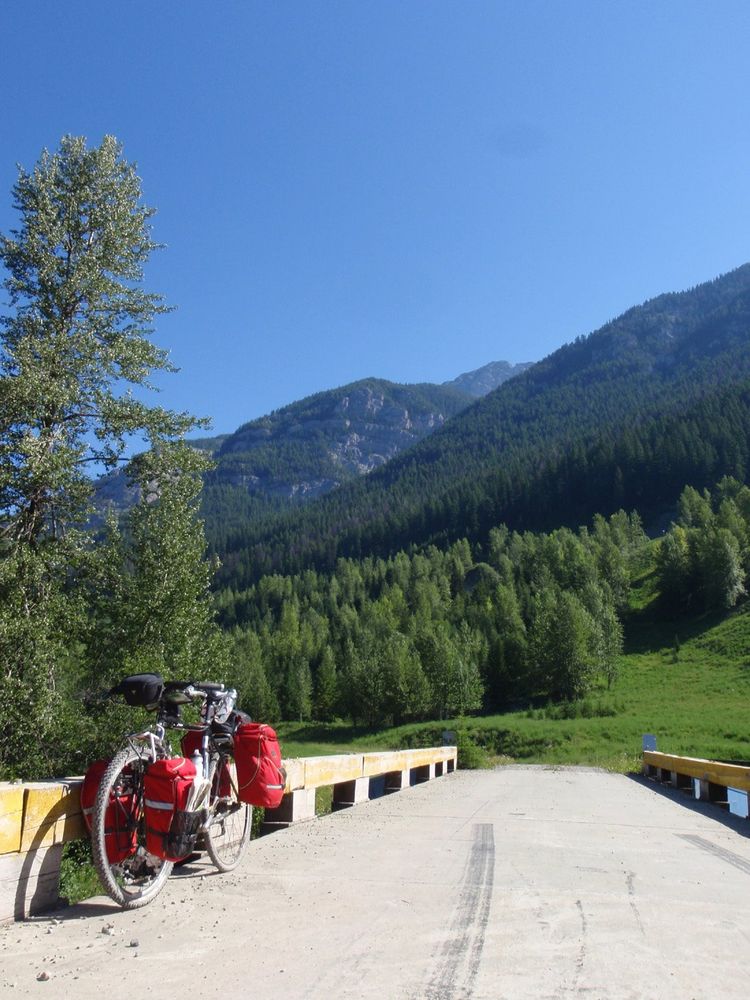 A bike fully loaded with camping gear sits on the side of a bridge in a beautiful, lush green valley at the base of some mountains with a clear blue sky above.