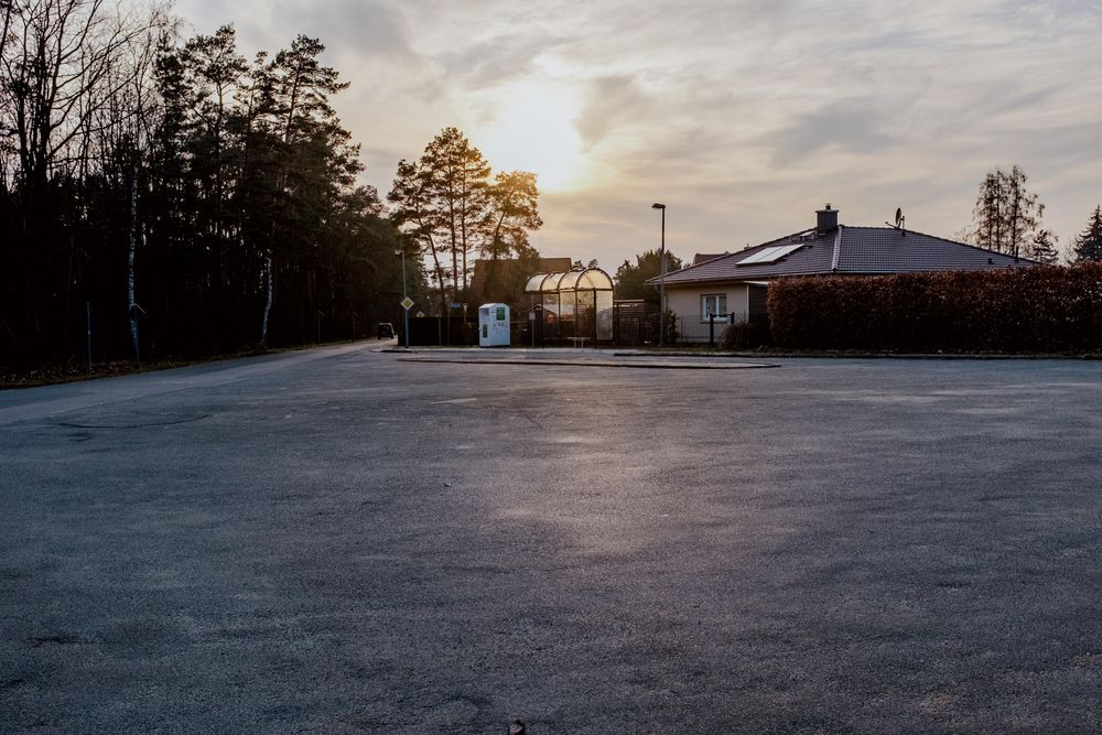 Das Bild zeigt eine große Fläche Asphalt, in der Bildmitte hinter der Fläche steht ein bushäuschen, größtenteils aus Glas, in dem die Abendsonne schimmert 