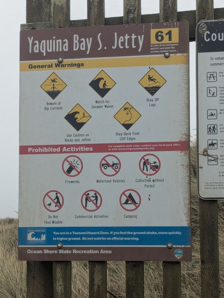 Sign that reads "Yaquina Bay S. Jetty" with several images of dangerous scenenarios: a swimmer crossing a rip current, a swimmer being hit from behind by a sneaker wave, a person falling off a log into the ocean, a person on a jetty about to get taken over by a large wave, and a person falling off a rocky cliff. 