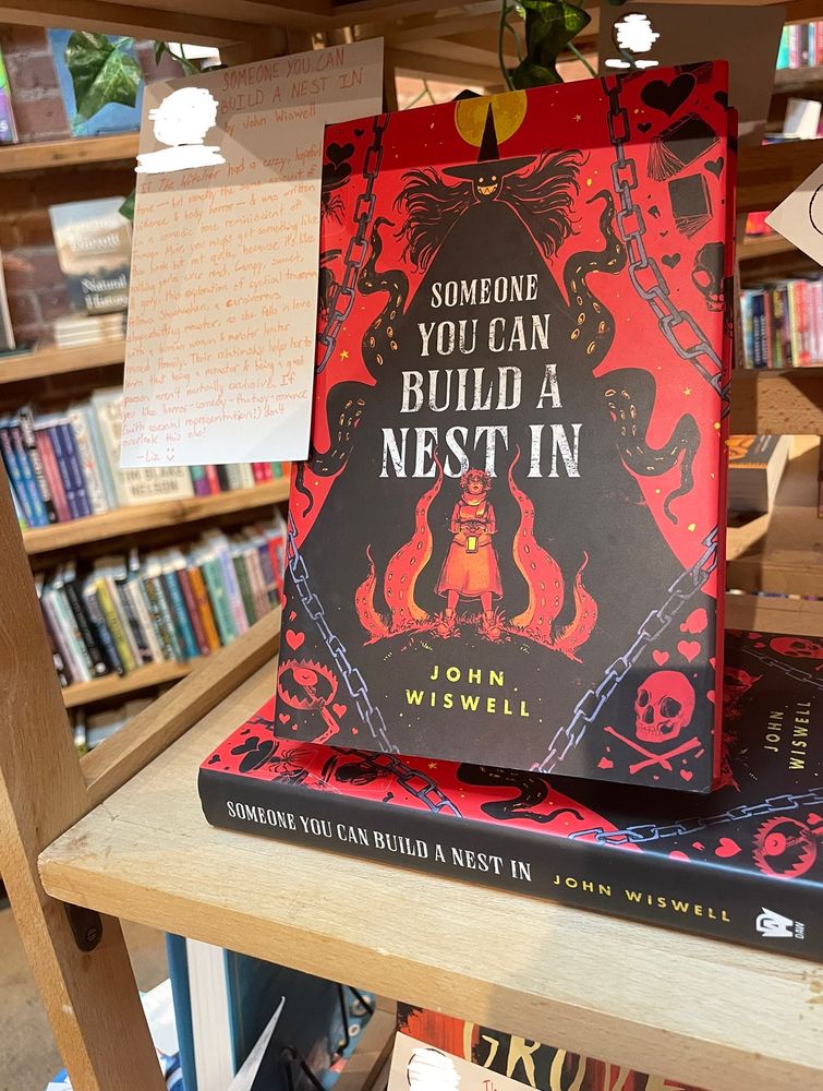 a picture of the cover of the book ‘someone you can build a nest in’ on the shelf at a bookstore. the cover features a woman holding a lantern as a smiling, inhuman silhouette looms behind her. a handwritten recommendation is attached to the book.