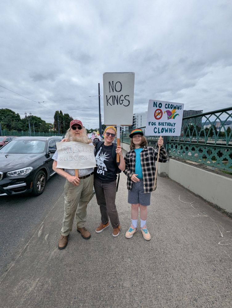 Three protesters on a sidewalk holding signs. They read "rebuild democracy", "no Kings", and "no crowns for birthday clowns"