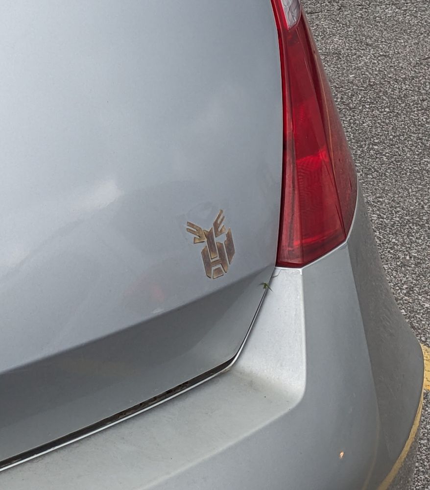 Deteriorated Autobot insignia on the back of a silver sedan. Looks like an old man with bushy eyebrows 