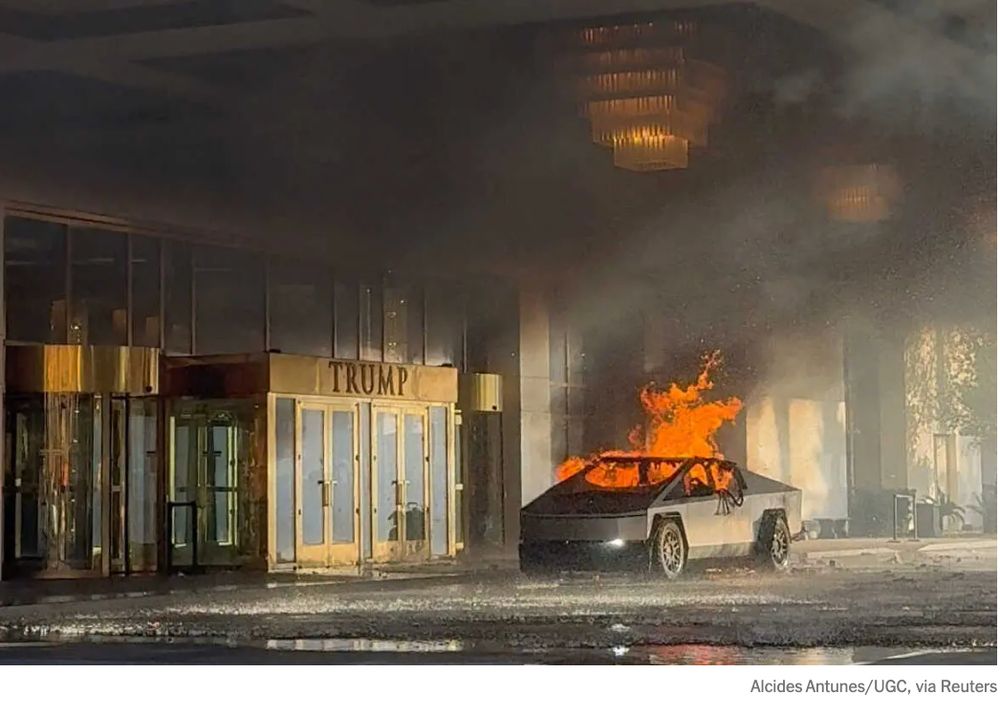 A medium-long-distance photograph of a burning Tesla Cybertruck parked just outside the doors of a Trump hotel. Sunlight is angling into the darkened area from the left. The flames are orange and clearly defined, sitting squarely in the truck and leaping through the roof and the window, giving off a vile-looking near-black smoke that veils the squarish tiers of the outdoor chandeliers on the canopy above. Above the brass-framed pair of glass double doors is more brass, in a panel with raised brass letters reading TRUMP. The photo credit reads Alcides Antunes/UGC, via Reuters.