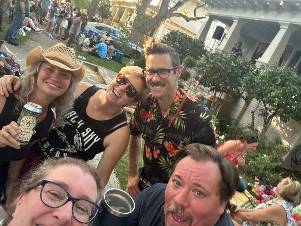 A group of five friends smiles in front of a band playing on a neighborhood porch