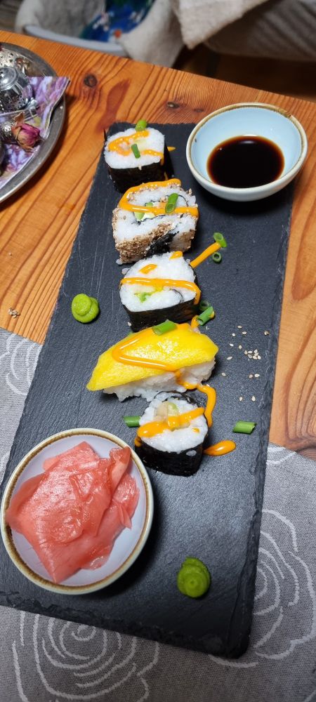 Ein heller Holztisch mit einem graugemusterten Deckchen. Darauf eine schwarze Platte mit zwei hellblauen Schalen, eine gefüllt mit rosa eingelegtem Ingwer, eine mit Sojasoße. Außerdem liegen 5 Sushistückchen auf der Platte, drei mit dunkler Alge außen, eine ohne Alge mit einem Mangostreifen, eine mit der Alge innen und Sesamkörnern außen. Orangene Soße ist darüber verteilt, ebenso ein paar Lauchzwiebelröllchen und Wazabitupfen