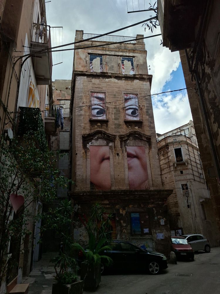 Un immeuble vétuste de la Città vecchia dont les fenêtres sont habitées par des yeux et un bas de visage humains 