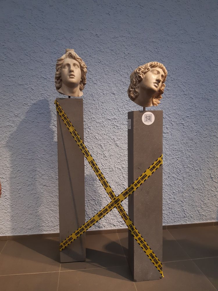 Deux têtes en marbre antiques exposées sur des piliers devant un mur.  A gauche celle d'un guerrier avec un casque et les yeux levés au ciel. A droite celle, affaissée sur le côté d'une femme, yeux clos. Des rubans jaunes et noirs barrent l'accès aux piliers.