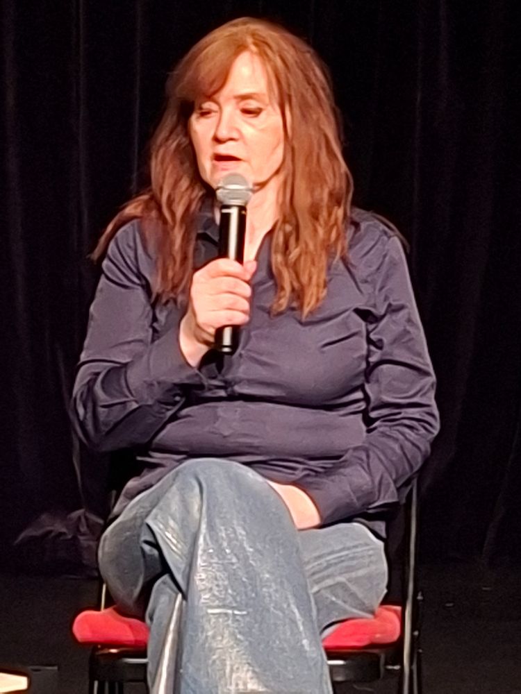 Photo rapprochée de l'écrivaine assise sur une chaise et parlant dans un micro. Elle a de longs cheveux roux et est venue d'une chemise bleu foncè et d'un jean argenté 