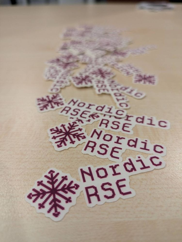 Picture of Nordic-RSE stickers (snowflake and the term Nordic-RSE in purple pixel style) on a table 