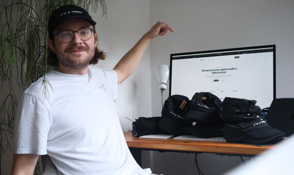 me, wearing a cap with « I’m a teapot » written - next to the display showing the openstatus website and a pile of the same caps on the keyboard.