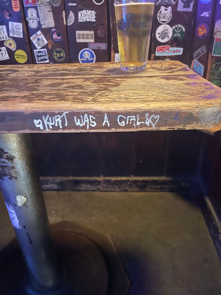 A grafito on the edge of a table in a sticker-covered bar reading, “Kurt was a girl.”