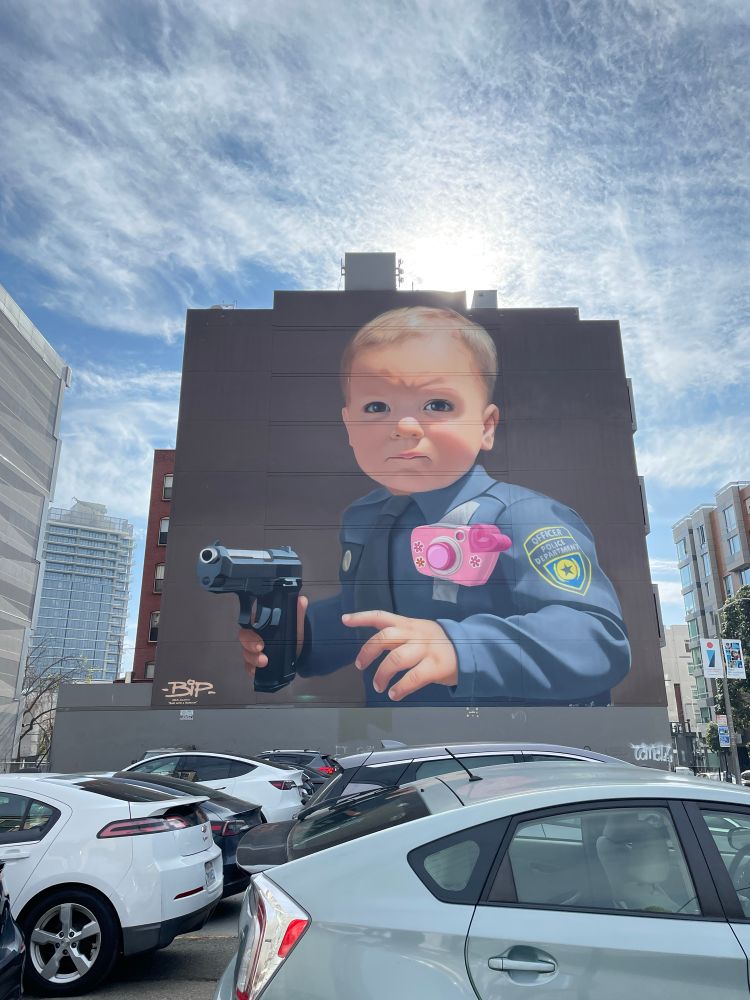 A four-story mural of a baby in a police uniform pointing a realistic gun. San Francisco.