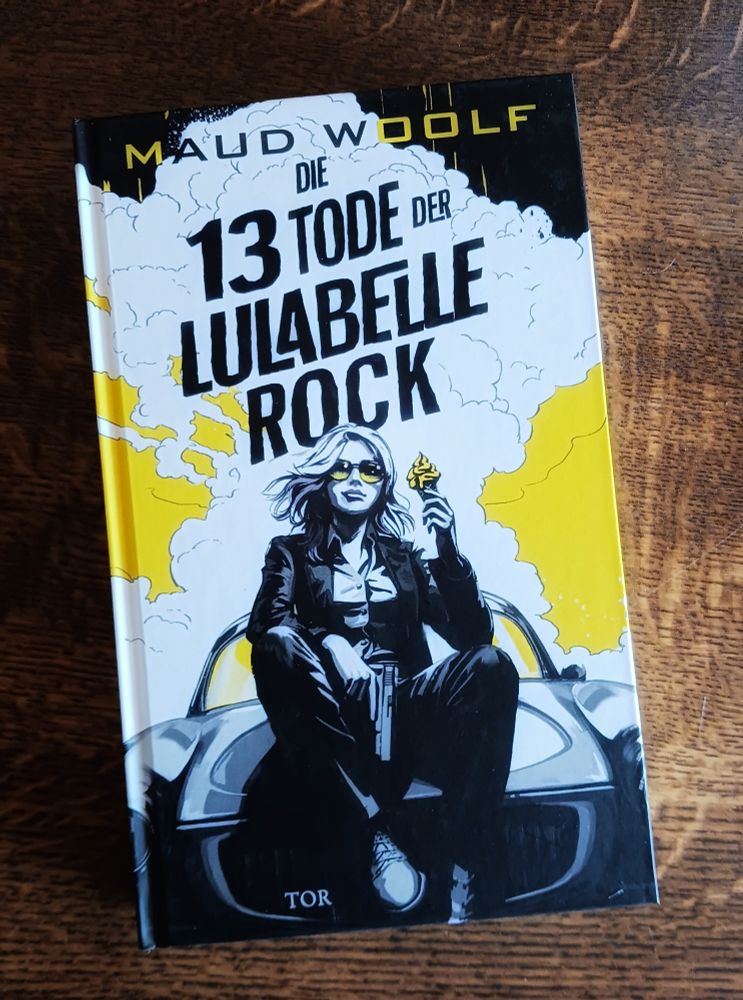 "Die 13 Tode der Lulabelle Rock" von Maud Woolf
Das Cover zeigt eine Comiczeichnung einer Frau, die mit einer Pistole in der rechten und einem Softeis in der linken Hand auf der Motorhaube eines Sportwagens sitzt. Im Hintergrund ziehen vor einem schwarzen Himmel und einem gelben Horizont weiße Wolken auf.