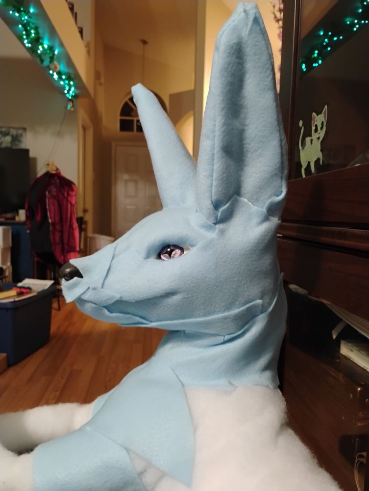Profile view of the left side of a large plush animal statue resembling a fox or a jackal. This will eventually become an almost life sized Glaceon who will lounge majestically on the shoulders of my dear husband. The Glaceon has one purple eye seen in this photo.