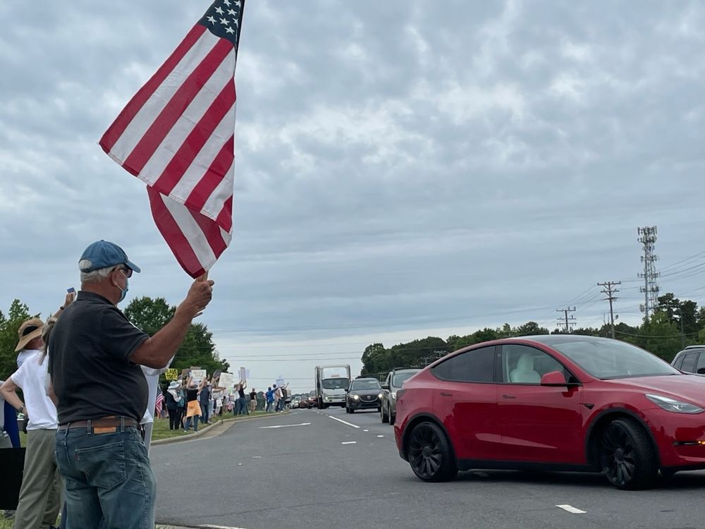 Line of Tesla/Musk protesters including one with American flag facing traffic and a red Tesla turning away