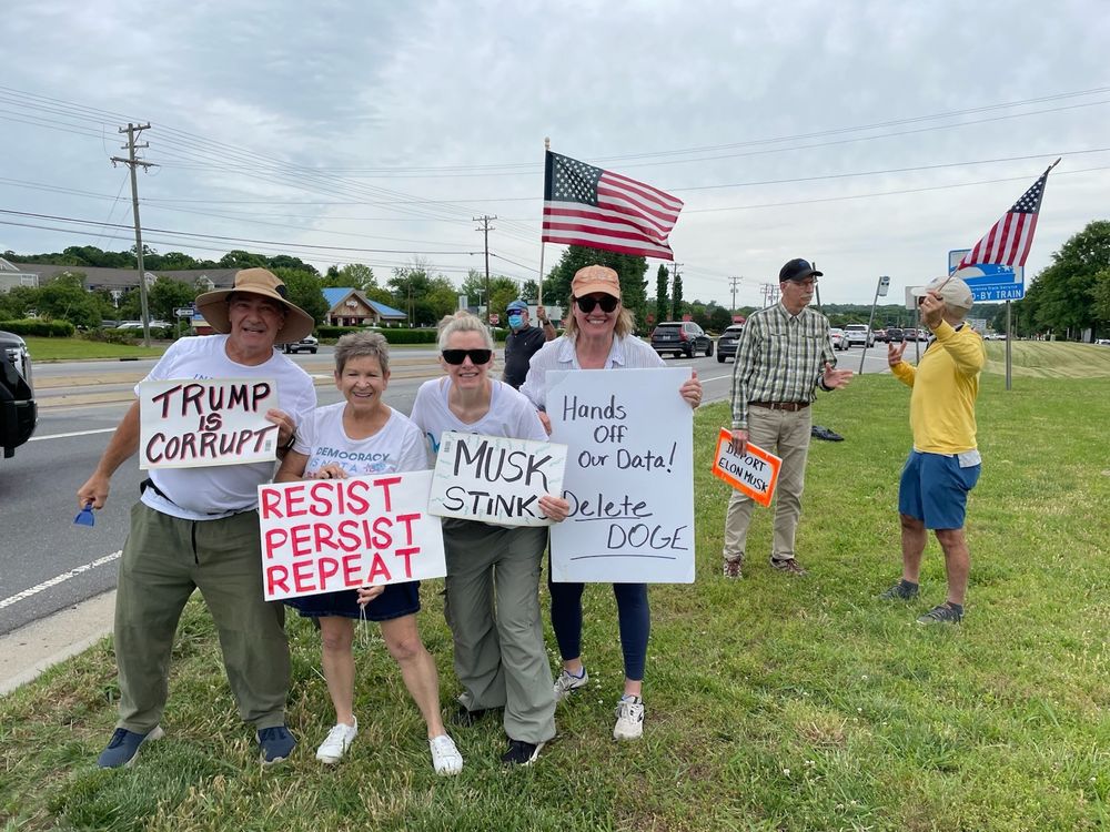 Tesla/Musk protesters with signs Trump is Corrupt, Resist Persist Repeat, Musk Stinks, Hands Off Our Data Delete Doge