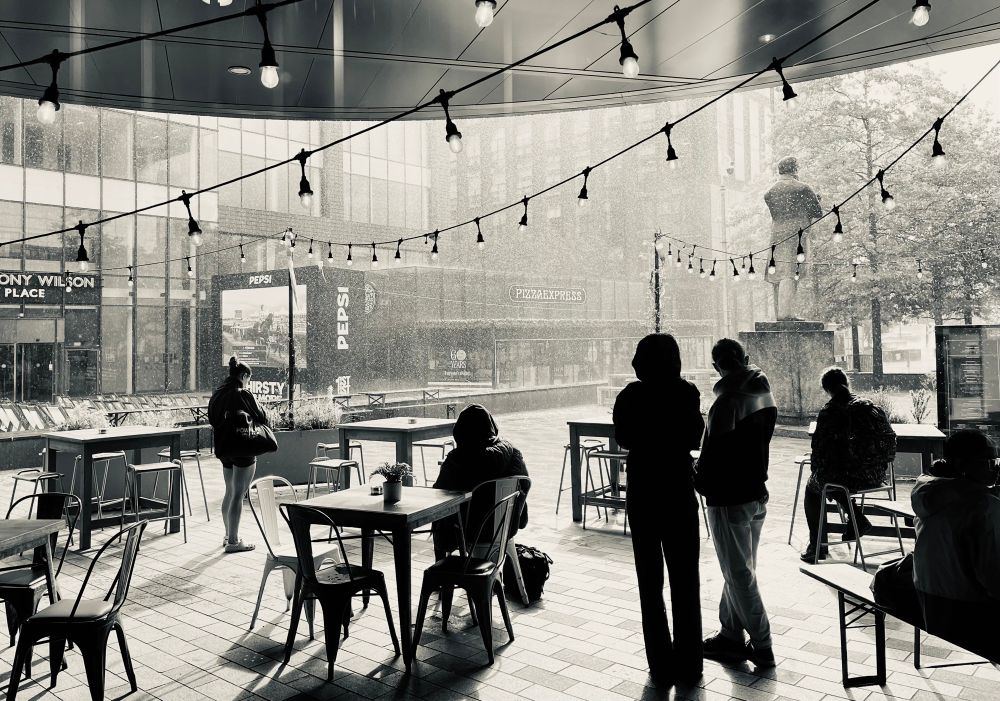Monochrome pic of a paved area with chairs & tables. A few people sitting or standing and looking out at the rain. Strings of lights above them & buildings in the background. A statue in the near distance on the right 