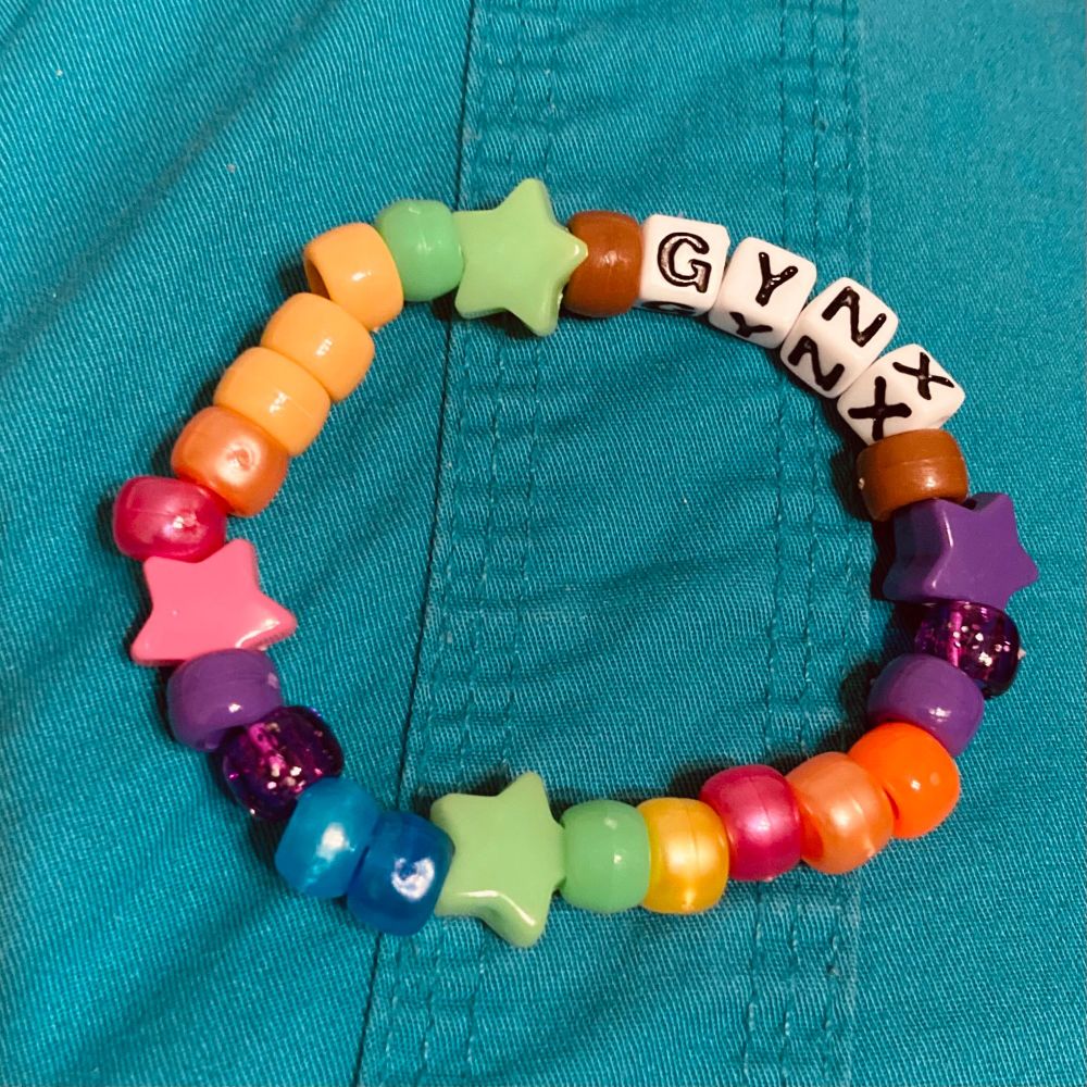 A colorful bracelet made of plastic beads; primarily normal pony beads with a few star-shaped beads, and 4 cubical letter beads which spell “GYNX” (the alias of a musician).