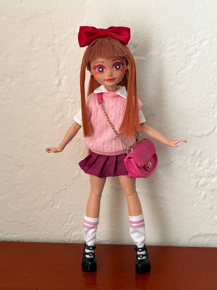 A custom doll based off of Blossom from The Powerpuff Girls. She wears a pink sweater vest with a white short shleeved dress shirt, a dark pink skirt, white socks with two pink stripes with black shoes. She has a pink crossbody bag. Her hair is done up in a ponytail and she has a red bow on top of her head. 