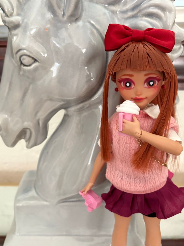Another picture of the Blossom doll, she's holding a camera in one hand and a smoothie in the other hand. She's in front of a bust of a horse.