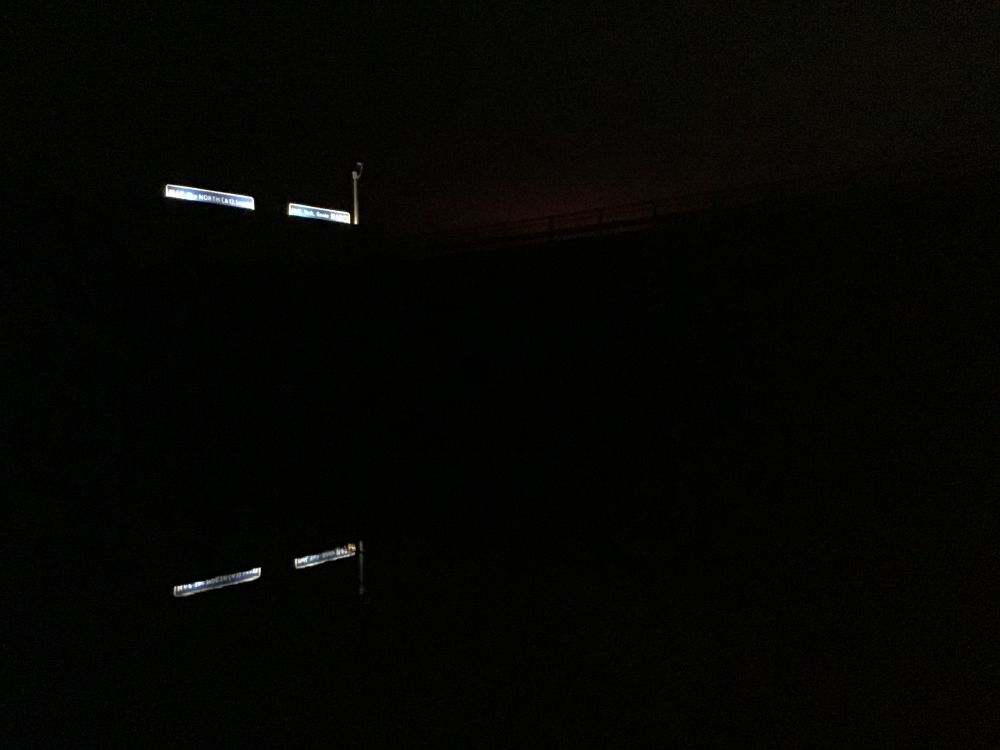 An almost pitch-dark photograph in which only the gantry of the M18, at its intersection with the Aire and Calder Navigation and the Dutch River, is visible (above the motorway, and reflected on the water).