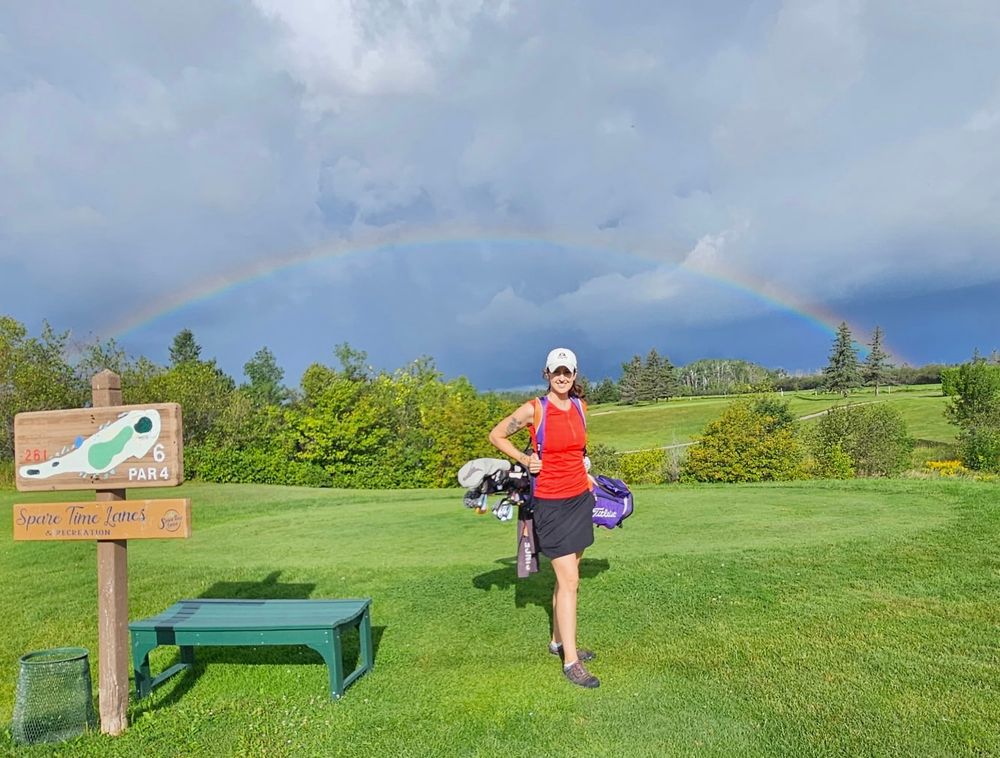 I am wearing golf attire and carrying a golf bag while standing on a tee box. There is a lush green landscape and a full rainbow behind me.