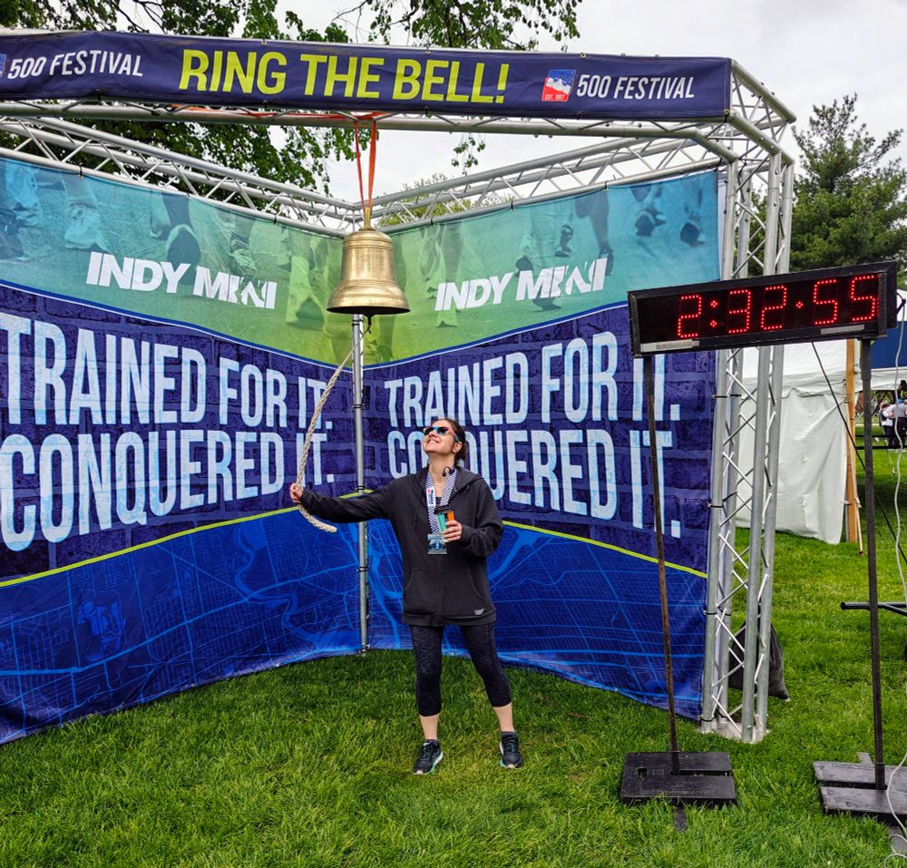 Martine ringing the personal record bell at the Indi Mini festival after finishing her half marathon in 2:32:55. She is wearing athletic gear while holding her coffee cup.