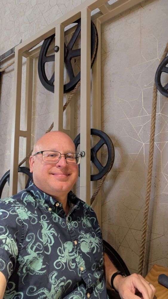 Me, in a blue shirt decorated with octopi, next to a wall. On the wall is a frame onto which are mounted several large pulleys with rope running between them. The rope is connected to a large door. 

This building was used for several industrial and warehousing businesses in the past. 