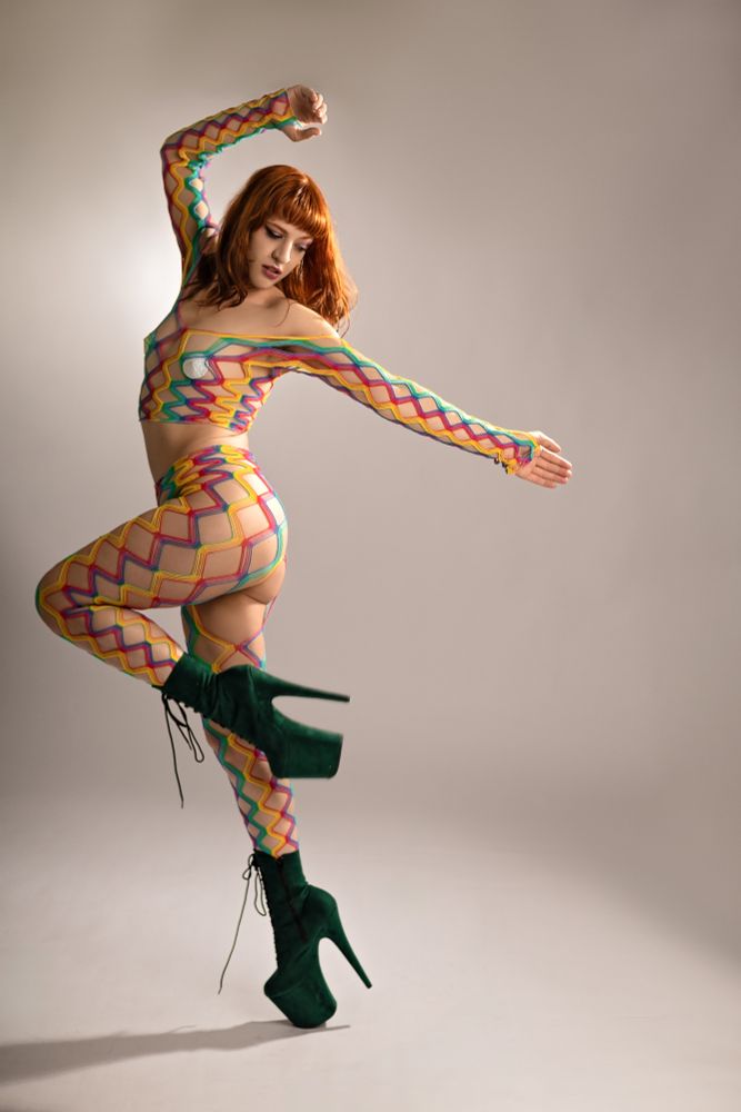 Friday poses in a white studio, wearing a rainbow fishnet two piece in leggings and a long sleeve crop top, with large 8 inch emerald green Pleaser boots. They are standing on one foot, slightly reminiscent of a sexy flamingo with their other leg bent at the knee. One arm is bent over their head and the other extended behind them, frozen in motion