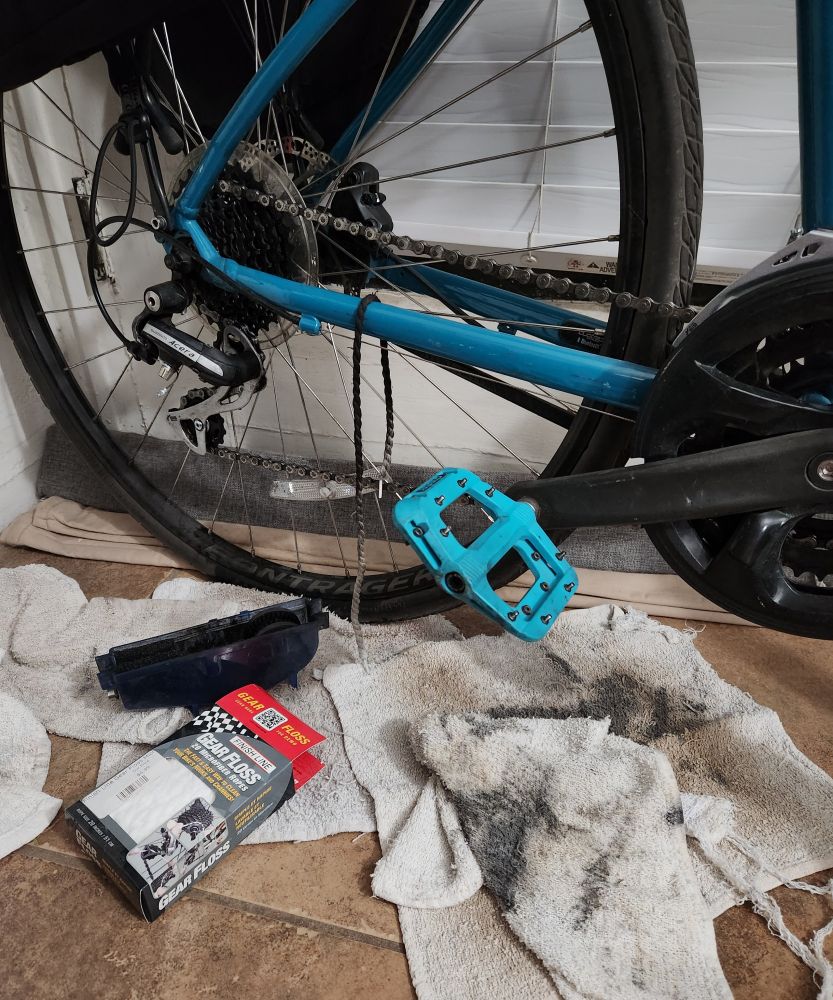 Photo of the rear tire of a bike with shop rags on the floor beneath. Also shown, chain cleaning gadget on the floor and used gear floss draped over the bike frame. 