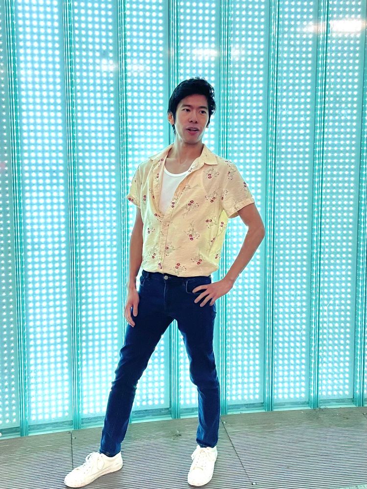 Man wearing a short sleeve button-down shirt patterned with small red flowers layered over a white tank top, jeans, and white sneakers