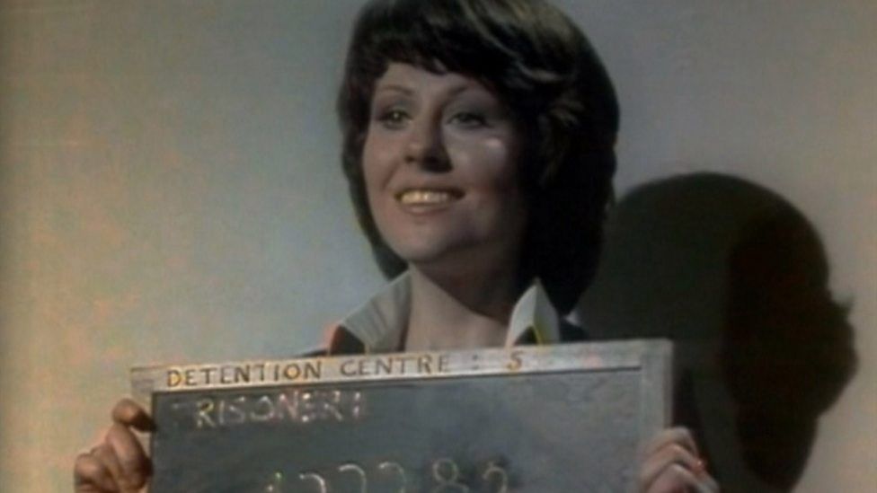 Sarah Jane Smith (Elisabeth Sladen) smiling as her mugshot is taken in Part 1 of Doctor Who: Invasion of the Dinosaurs (1974)