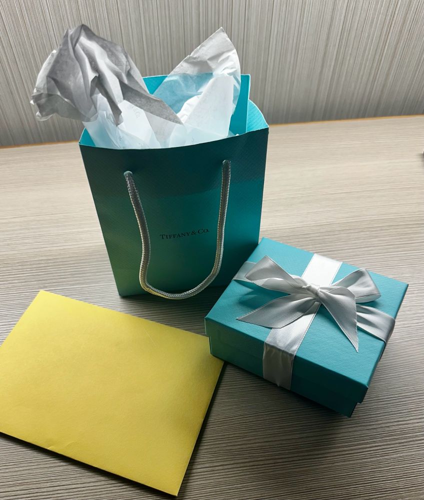 A Tiffany blue gift bag and matching box tied with a white bow sitting beside a yellow envelope.