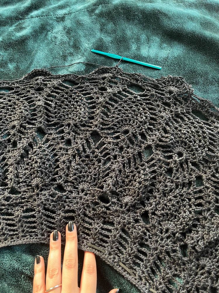 black lace-like crochet in a repeating pattern that resembles big acorns or leaves. a hand is at the bottom and a crochet hook rests at the top, to show the project is fairly large and getting larger.