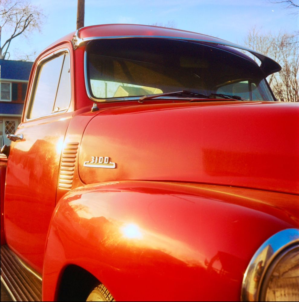 A vintage Chevrolet 3100 pickup truck in glossy red paint, reflecting the sunlight. The image focuses on the front fender, hood, and driver’s side door, with the ‘3100’ emblem prominently displayed. The truck features a distinctive sun visor over the windshield. The reflection of the sky and surroundings is visible on the truck’s curved surfaces. A suburban house with blue siding and white trim is partially visible in the background, along with bare trees under a clear blue sky.