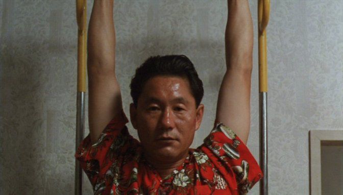 Takeshi Kitano et sa tête merveilleusement impassible, suspendu par les bras à une barre. Il porte une chemise rouge à motifs. Si cette photo ne le montre pas, il est en réalité en slip.
Tiré de son film "Jugatsu".