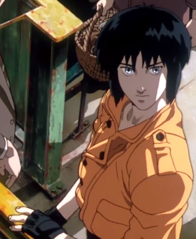 Motoko Kusanagi, dans le Ghost in the Shell de Mamoru Oshii. Elle porte un blouson jaune avec une fermeture à boutons et col façon imperméable, des mitaines noires au main. Elle s’appuie sur une rambarde et regarde légèrement vers le haut en direction de l’objectif.