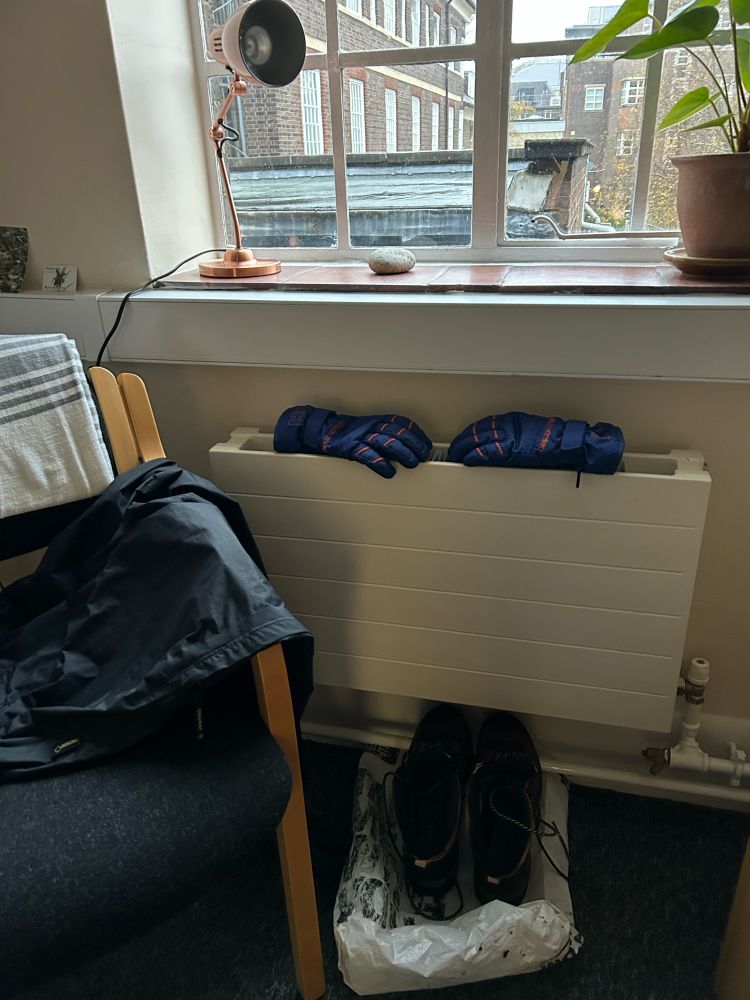 Wet weather gear drying around a radiator beneath a window. Outside it is grey and raining hard.