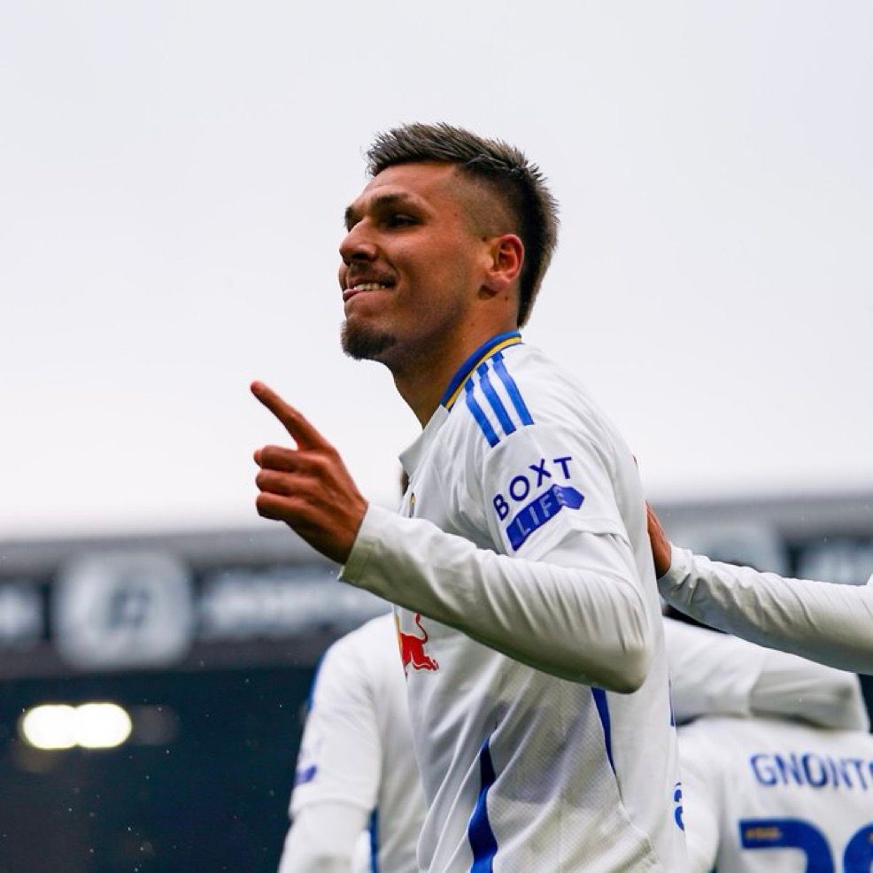 Joel Piroe celebrates after scoring a goal, pointing upward with his right hand while smiling. He is wearing a white Leeds United kit with blue and yellow accents, and the sponsor “BOXT” visible on his sleeve. Teammates, including one with “Gnonto” on the back of his jersey, gather behind him.