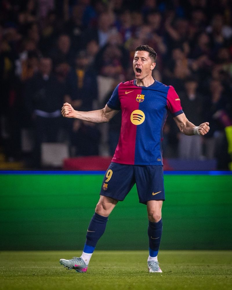 Robert Lewandowski, wearing the red and blue FC Barcelona home kit with the number 9, celebrates passionately on the pitch with clenched fists and an open-mouthed roar. He stands in front of a bright LED advertising board, with a blurred crowd cheering in the background.