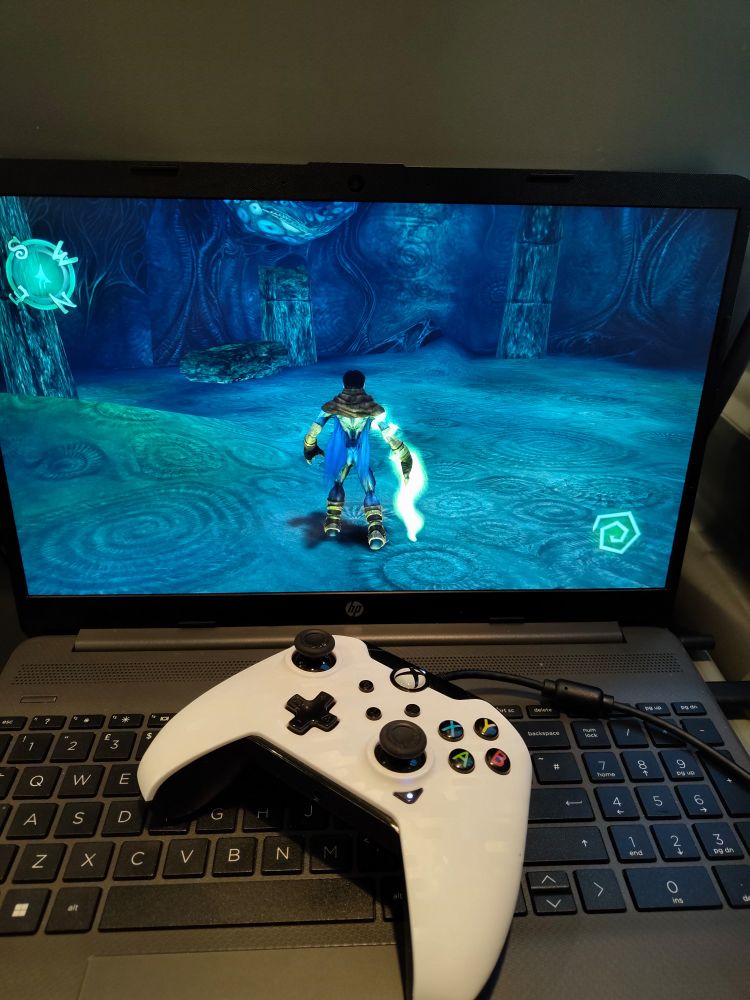 A grey laptop with a white Xbox controller resting on the keyboard. The screen shows a still image from the game Soul Reaver 1 